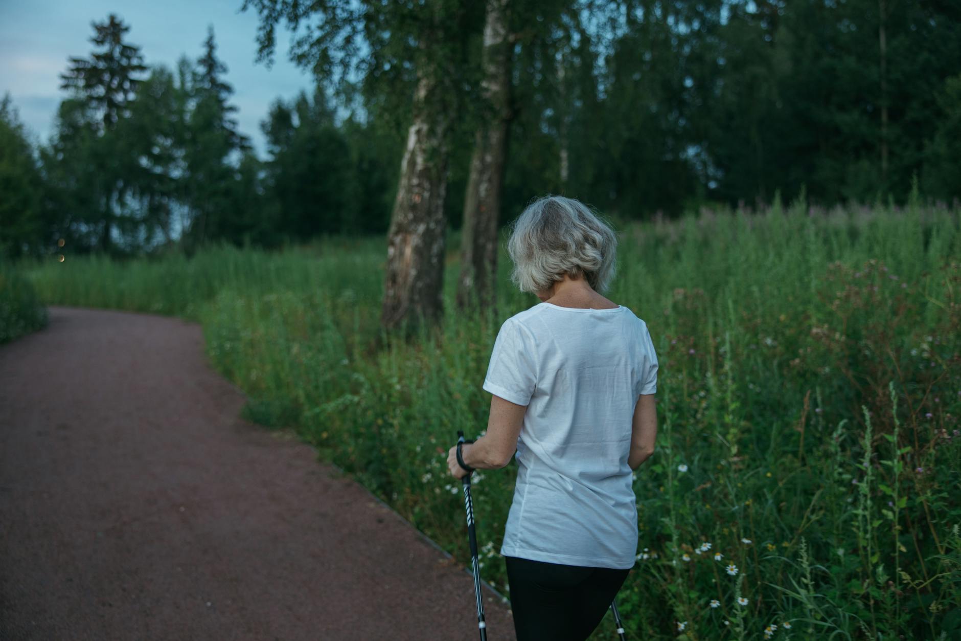 Starsza kobieta z kijkami nordic walking idąca alejką w parku