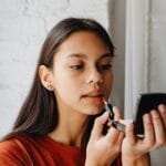 Młoda kobieta nakłada tint na usta, patrząc w lusterko przy oknie