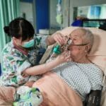 Pielęgniarka zakłada starszemu pacjentowi maskę tlenową w szpitalnym łóżku